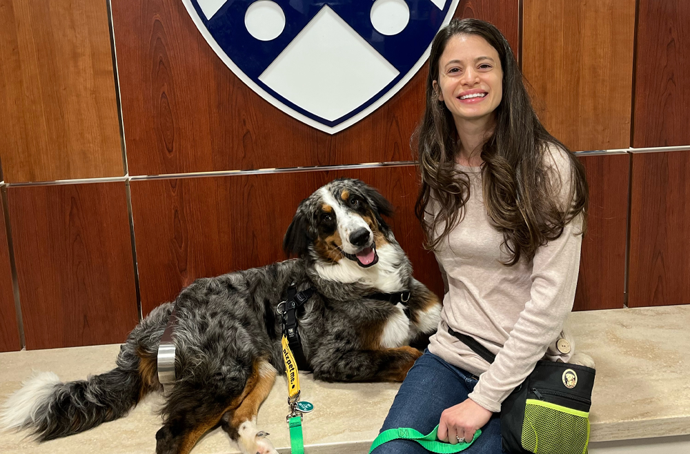 Jen Abdul-Hakeem sits with Marley on a bench with the Penn shield on a wall behind them.